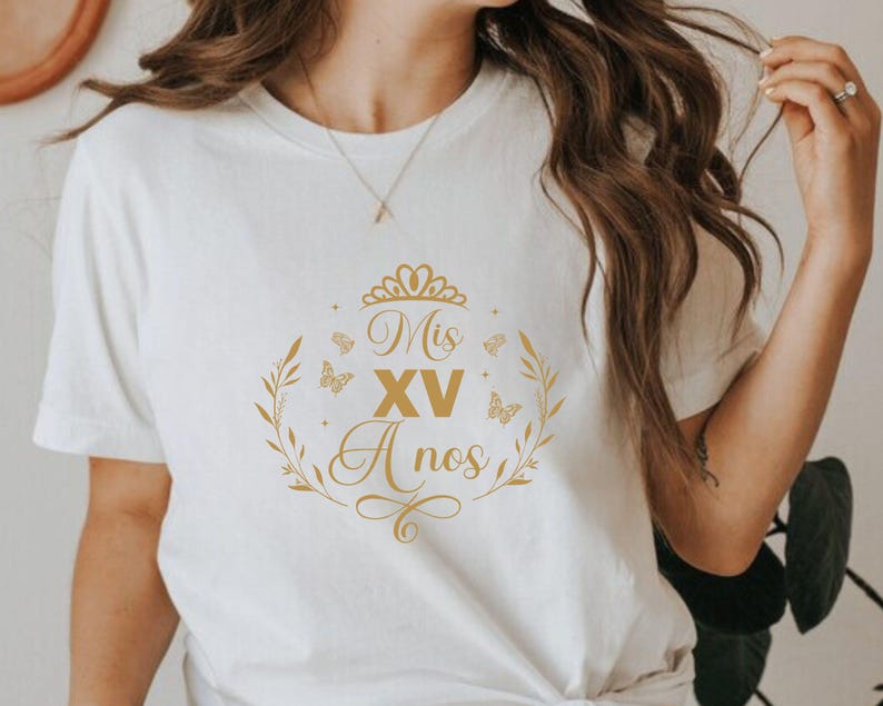 Op de afbeelding: Wit T-shirt met een gouden grafisch ontwerp met de tekst "Mis XV A&ntilde;os" met een kroon en vlinders.