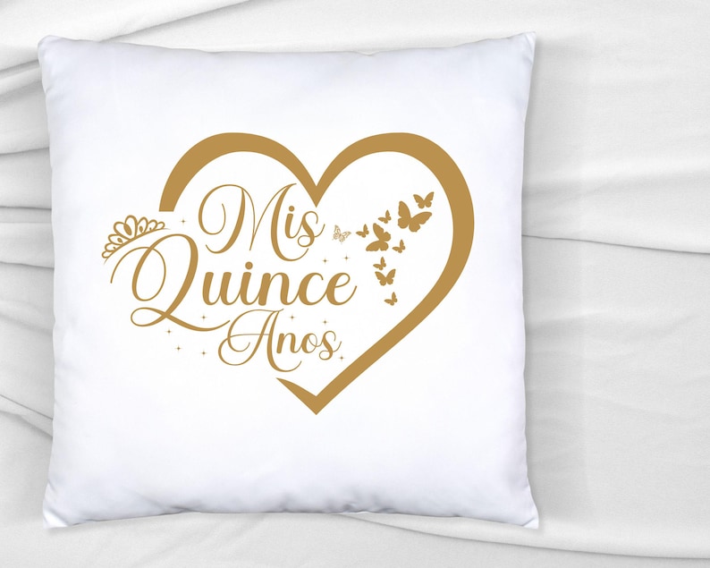 Op de afbeelding: Wit kussen met een gouden hartvormig ontwerp met de tekst "Mis Quince Anos" en gouden vlinders rond het hart.