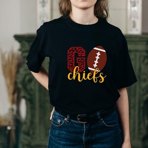 May include: Black t-shirt with a red and white "GO" graphic with a leopard print pattern. The "O" is replaced with a brown and white football. The text "chiefs" is in yellow below the graphic.