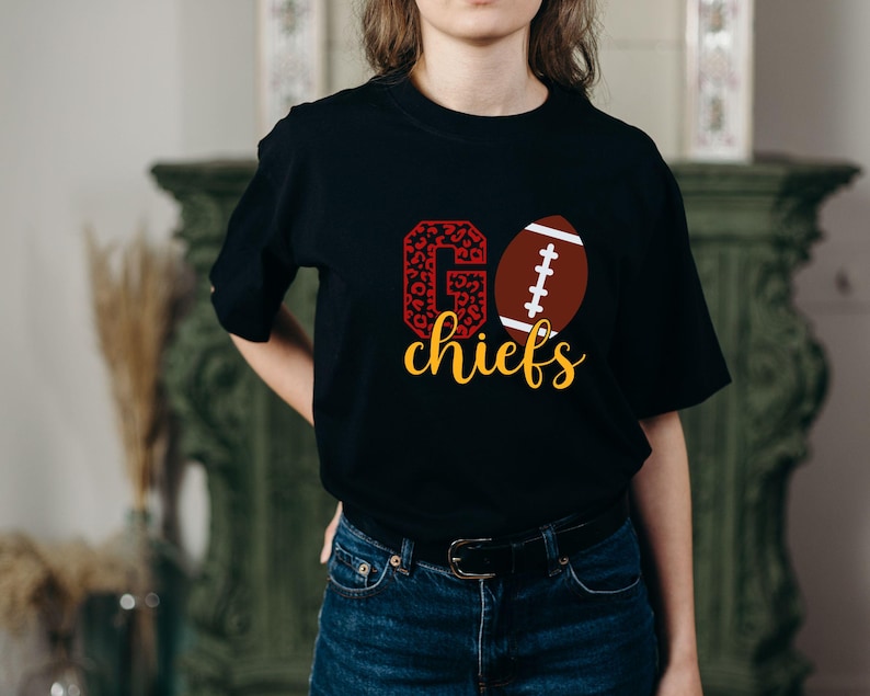 May include: Black t-shirt with a red and white "GO" graphic with a leopard print pattern. The "O" is replaced with a brown and white football. The text "chiefs" is in yellow below the graphic.