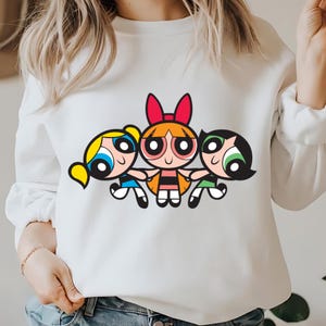 May include: White sweatshirt with a graphic print of the Powerpuff Girls, Blossom, Bubbles, and Buttercup. The girls are cartoon characters with colorful hair and outfits.