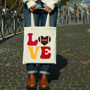 May include: White tote bag with the word "LOVE" in red, yellow, and brown letters. A brown football in a heart shape is above the "O".