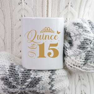 Op de afbeelding: Een witte keramische mok met een gouden ontwerp met de tekst "Mis Quince 15" met een kroon en een vlinder. De mok wordt vastgehouden door een persoon die een witte trui draagt.
