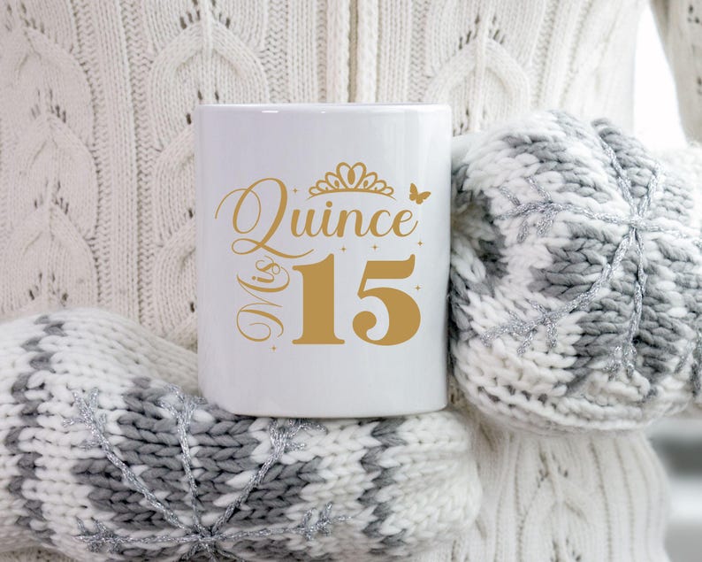 Op de afbeelding: Een witte keramische mok met een gouden ontwerp met de tekst "Mis Quince 15" met een kroon en een vlinder. De mok wordt vastgehouden door een persoon die een witte trui draagt.