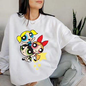May include: White sweatshirt with a graphic print of the Powerpuff Girls, Blossom, Bubbles, and Buttercup, in a playful pose. The graphic is in bright colors and features a yellow star.