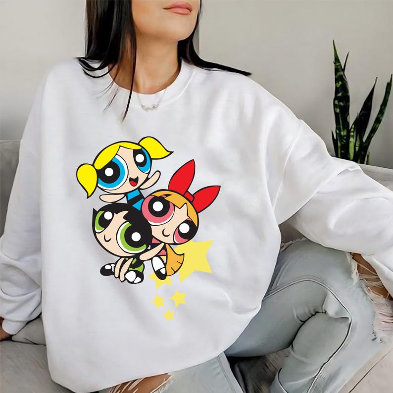 May include: White sweatshirt with a graphic print of the Powerpuff Girls, Blossom, Bubbles, and Buttercup, in a playful pose. The graphic is in bright colors and features a yellow star.
