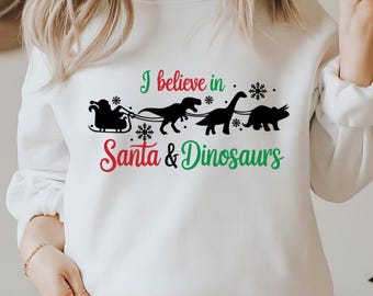 May include: White sweatshirt with a Christmas-themed graphic that says "I believe in Santa & Dinosaurs". The graphic features a Santa Claus in a sleigh pulled by three dinosaurs.
