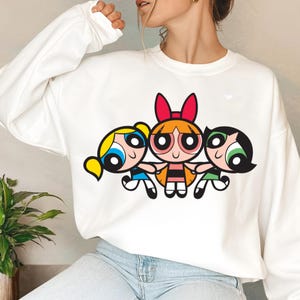 May include: White sweatshirt with a graphic print of the Powerpuff Girls cartoon characters. The characters are Bubbles, Blossom, and Buttercup. The characters are in a line, holding hands, and smiling.