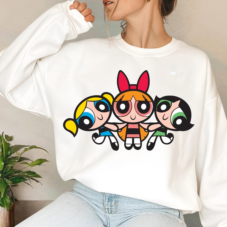 May include: White sweatshirt with a graphic print of the Powerpuff Girls cartoon characters. The characters are Bubbles, Blossom, and Buttercup. The characters are in a line, holding hands, and smiling.
