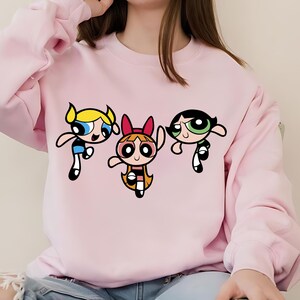 May include: Pink sweatshirt with a graphic print of the Powerpuff Girls cartoon characters. The characters are Bubbles, Blossom, and Buttercup.