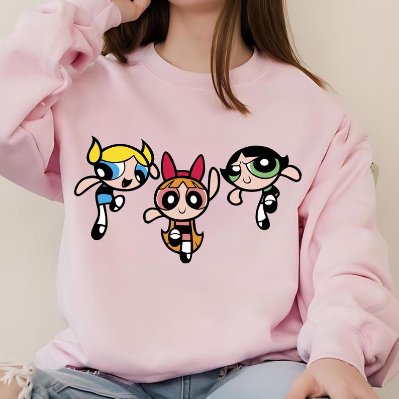 May include: Pink sweatshirt with a graphic print of the Powerpuff Girls cartoon characters. The characters are Bubbles, Blossom, and Buttercup.