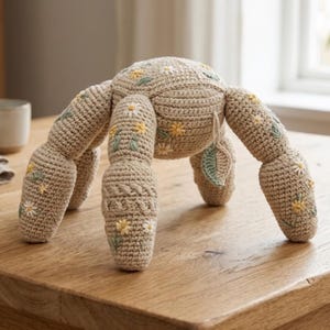 Puede incluir: Un animal de peluche tejido a ganchillo en color beige con cuerpo redondeado y cuatro patas, decorado con flores de margarita amarillas y blancas bordadas y hojas verdes. La superficie de hilo texturizado incluye una pequeña hoja colgante.