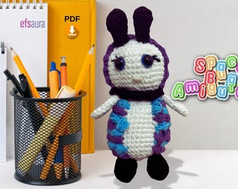 May include: A crocheted purple and blue bug amigurumi toy with black eyes and a white face. The toy is standing on a white surface with a yellow binder and a black pencil holder in the background. The text "Space Bug Amigurumi" is in the background.