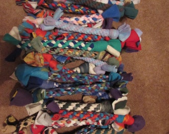 May include: A collection of colorful, braided dog toys made from various fabrics. The toys feature a variety of patterns and colors, including blue, red, green, and patterned fabrics. Each toy is knotted at both ends.