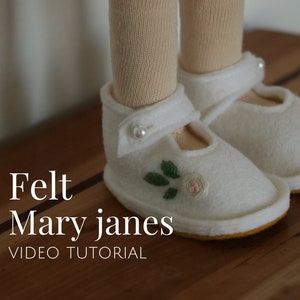 Puede incluir: Zapatos Mary Jane de fieltro blanco con ribete amarillo y una pequeña flor blanca bordada en la punta. Los zapatos están sobre una superficie de madera.