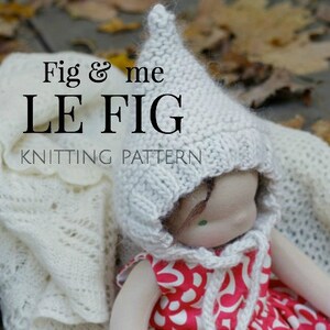 K&ouml;nnte beinhalten: Eine gestrickte Puppe, die einen wei&szlig;en H&auml;kelhut und ein rot-wei&szlig;es Blumenkleid tr&auml;gt. Die Puppe sitzt auf einer wei&szlig;en H&auml;keldecke mit braunen Bl&auml;ttern im Hintergrund. Der Text "Fig & me LE FIG KNITTING PATTERN" ist im Bild sichtbar.