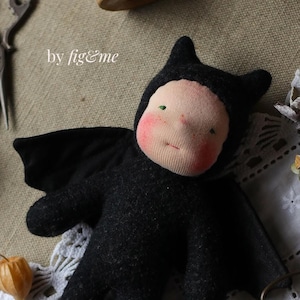May include: A black felt bat costume with wings for a doll. The doll has a pink face with green eyes and a stitched mouth. The text "by fig&me" is visible in the image.