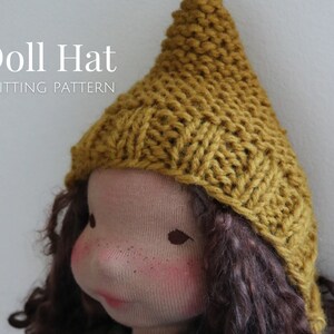 K&ouml;nnte beinhalten: Eine gestrickte Puppenm&uuml;tze in senfgelber Farbe. Die M&uuml;tze ist mit einem einfachen Stichmuster gearbeitet und hat eine spitze Krone.