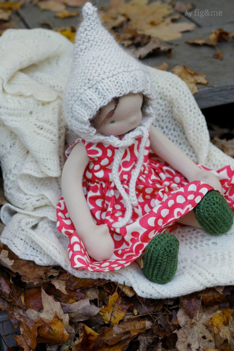 K&ouml;nnte beinhalten: Eine handgefertigte Puppe mit einer wei&szlig;en Strickm&uuml;tze und einem rot-wei&szlig; gemusterten Kleid. Die Puppe sitzt auf einer wei&szlig;en H&auml;keldecke mit gr&uuml;nen Strickschuhen.