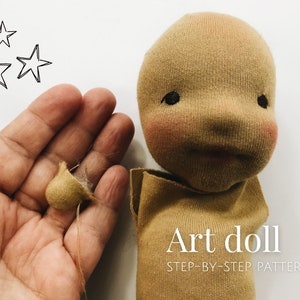 May include: A close-up of a beige fabric art doll head with black eyes and a stitched-on mouth. The doll's head is being held in a hand, and a small beige fabric ball is in the palm of the hand. The text "Art doll" and "STEP-BY-STEP PATTERN" is visible below the doll's head.