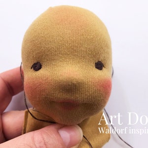 May include: Close-up of a beige fabric doll head with embroidered eyes and a stitched mouth. The doll is being held by a hand. The text "Art Doll Waldorf inspired" is visible below the doll's head.