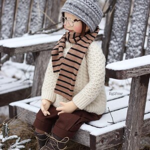 K&ouml;nnte beinhalten: Eine Puppe tr&auml;gt einen wei&szlig;en Pullover, einen braunen und wei&szlig;en gestreiften Schal, eine graue und wei&szlig;e Strickm&uuml;tze mit einem wei&szlig;en Bommel und braune Stiefel. Die Puppe sitzt auf einer mit Schnee bedeckten Holzbank.