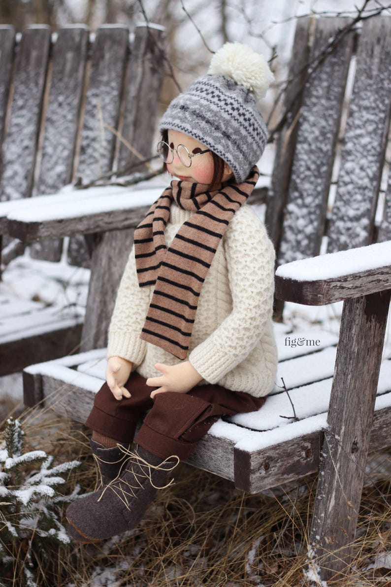 K&ouml;nnte beinhalten: Eine Puppe tr&auml;gt einen wei&szlig;en Pullover, einen braunen und wei&szlig;en gestreiften Schal, eine graue und wei&szlig;e Strickm&uuml;tze mit einem wei&szlig;en Bommel und braune Stiefel. Die Puppe sitzt auf einer mit Schnee bedeckten Holzbank.