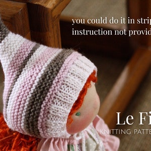 K&ouml;nnte beinhalten: Eine gestrickte Puppenm&uuml;tze mit rosa, wei&szlig;en und braunen Streifen. Die M&uuml;tze hat eine spitze Spitze und wird von einer Puppe mit roten Haaren getragen. Der Text "Le Fig Knitting Pattern" ist im Bild sichtbar.