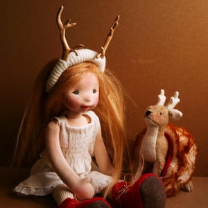 K&ouml;nnte beinhalten: Eine handgefertigte Puppe mit langen roten Haaren, die ein wei&szlig;es Kleid und ein Stirnband mit Geweih tr&auml;gt. Die Puppe sitzt mit einem kleinen Stofftier in Form eines Rehs.