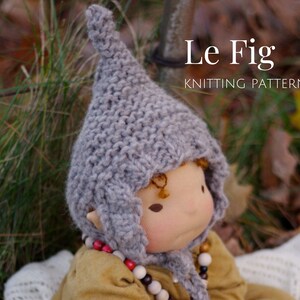 K&ouml;nnte beinhalten: Eine graue Strickm&uuml;tze mit einer spitzen Spitze, genannt "Le Fig Knitting Pattern", wird von einer Puppe mit einem hellbraunen Gesicht und braunen Augen getragen.
