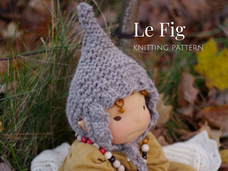K&ouml;nnte beinhalten: Eine graue Strickm&uuml;tze mit einer spitzen Spitze, genannt "Le Fig Knitting Pattern", wird von einer Puppe mit einem hellbraunen Gesicht und braunen Augen getragen.
