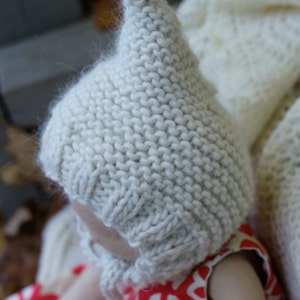 K&ouml;nnte beinhalten: Eine wei&szlig;e gestrickte Baby-M&uuml;tze mit einer spitzen Spitze. Die M&uuml;tze wird von einem Baby getragen, das ein rot-wei&szlig; gemustertes Kleid tr&auml;gt.