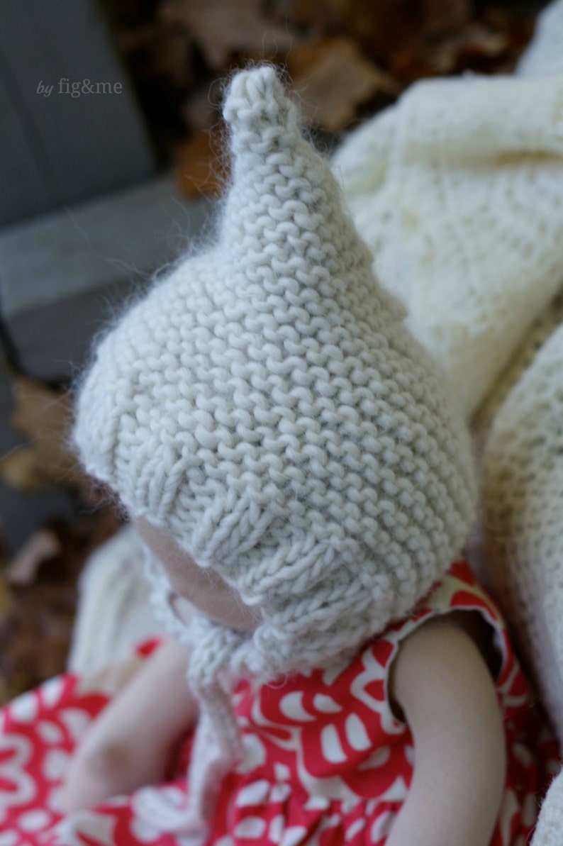 K&ouml;nnte beinhalten: Eine wei&szlig;e gestrickte Baby-M&uuml;tze mit einer spitzen Spitze. Die M&uuml;tze wird von einem Baby getragen, das ein rot-wei&szlig; gemustertes Kleid tr&auml;gt.