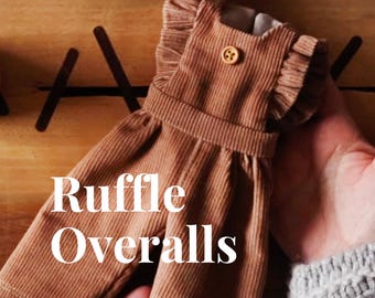 May include: Tan corduroy overalls featuring ruffled shoulder straps and a single small wooden button at the center of the chest. The text Ruffle Overalls is printed in white over the garment. The fabric has a distinct vertical ribbed texture.