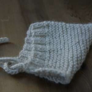 K&ouml;nnte beinhalten: Eine wei&szlig;e gestrickte Babyhaube mit einem einfachen Stichmuster. Die Haube hat eine Schleife, die unter dem Kinn gebunden werden kann.
