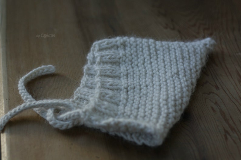 K&ouml;nnte beinhalten: Eine wei&szlig;e gestrickte Babyhaube mit einem einfachen Stichmuster. Die Haube hat eine Schleife, die unter dem Kinn gebunden werden kann.