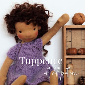 May include: A purple knitted dress with a lace pattern on a handmade doll with brown curly hair. The doll is holding up one arm and is standing next to a wooden shelf with a ruler on the side. The shelf has a walnut, acorns, and a wooden peg doll on it. The text "Tuppence art doll pattern" is written below the doll.