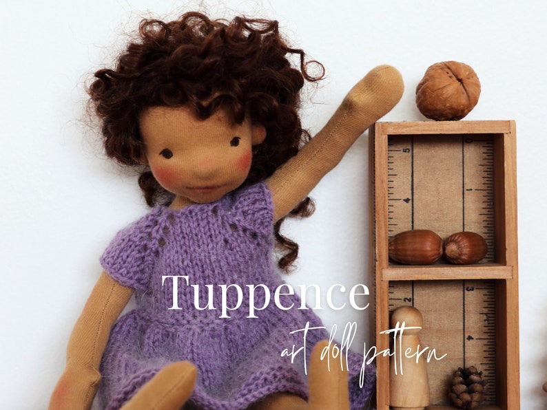May include: A purple knitted dress with a lace pattern on a handmade doll with brown curly hair. The doll is holding up one arm and is standing next to a wooden shelf with a ruler on the side. The shelf has a walnut, acorns, and a wooden peg doll on it. The text "Tuppence art doll pattern" is written below the doll.
