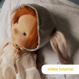 May include: A handmade doll with red hair wearing a gray hooded hat with bunny ears. The doll is wearing a white dress with a ribbon tied around the neck. The doll is lying on a white fabric surface. The text "video tutorial" is visible in the lower right corner of the image.