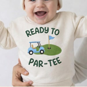 May include: A smiling baby wearing a cream-colored sweatshirt and a white beanie. The sweatshirt features a golf cart and the text "READY TO PAR-TEE" in green and blue. The baby is being held by someone.