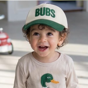 May include: A child wearing a beige long-sleeved shirt with an embroidered duck design, khaki shorts, and a cream-colored baseball cap with the word "BUBS" in green. The child is smiling. A red toy fire truck is in the background.