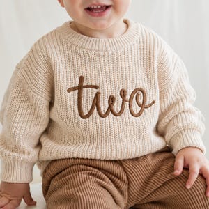 May include: A beige long-sleeved knit sweater featuring the word two embroidered in brown cursive script across the chest. The sweater has a ribbed crew neckline and ribbed cuffs, paired with brown corduroy pants with a vertical ribbed texture.