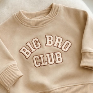 May include: A tan-colored sweatshirt with the words "BIG BRO CLUB" embroidered in a brown and white collegiate style. The sweatshirt has a crew neck and ribbed cuffs and hem. The garment is made of a soft material.