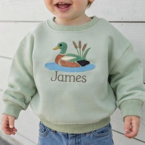 Puede incluir: Sudadera verde claro con un diseño de pato bordado en la parte delantera. El pato está en un estanque con juncos. El nombre "James" está bordado debajo. La sudadera tiene puños y dobladillo de canalé.