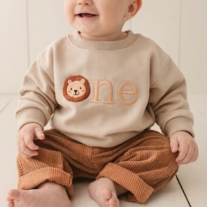 May include: A baby in a beige sweatshirt and brown corduroy trousers. The sweatshirt features a lion face and the word "one". The baby is sitting on a white wooden surface.
