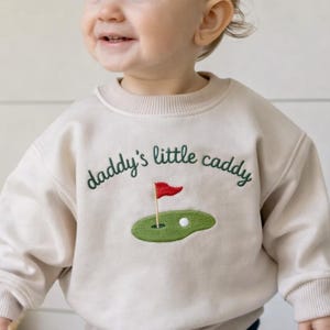 May include: A beige sweatshirt with the embroidered text "daddy's little caddy" in green, featuring a golf course design with a red flag and a golf ball. The child is smiling.