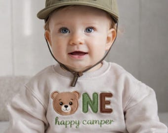 Puede incluir: Un bebé con una gorra de béisbol verde caqui con el texto "HAPPY CAMPER" y un parche con un gráfico de árbol. El bebé también lleva una sudadera beige con un gráfico de oso y el texto "ONE happy camper".