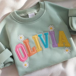 May include: A pale green sweatshirt featuring the name "OLIVIA" in embroidered, multi-coloured letters. The letters are outlined in a contrasting colour. Small daisy flower designs are embroidered around the name. The sleeves are cuffed.
