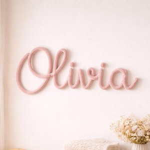 May include: A nursery scene with a pink knitted "Olivia" name sign on a white wall. A wooden chest of drawers displays folded blankets, a soft toy, and a vase of flowers. A wicker crib is in the foreground.