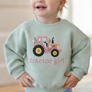 May include: A light sage green long-sleeve sweatshirt featuring an embroidered pink tractor with yellow wheels and floral accents. The text "tractor girl" is embroidered in pink lettering below the tractor design on the front of the garment.